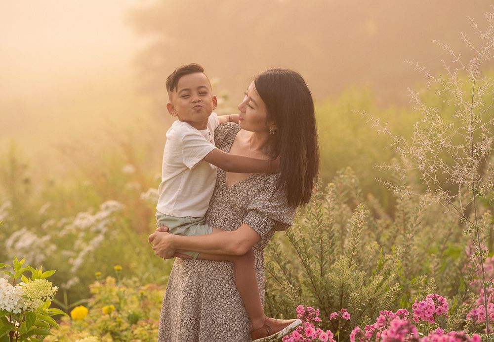 A mother and child share a tender moment in a garden at sunset surrounded by wildflowers and golden light.