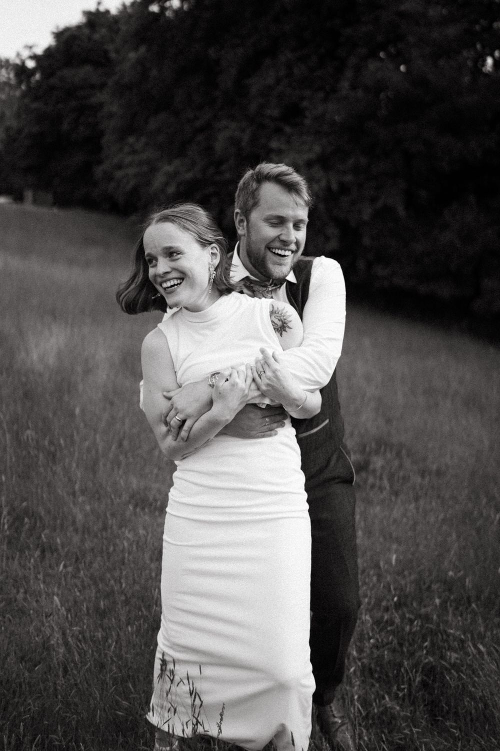 A couple embraces joyfully in a grassy field, with the scene captured in black and white photography.