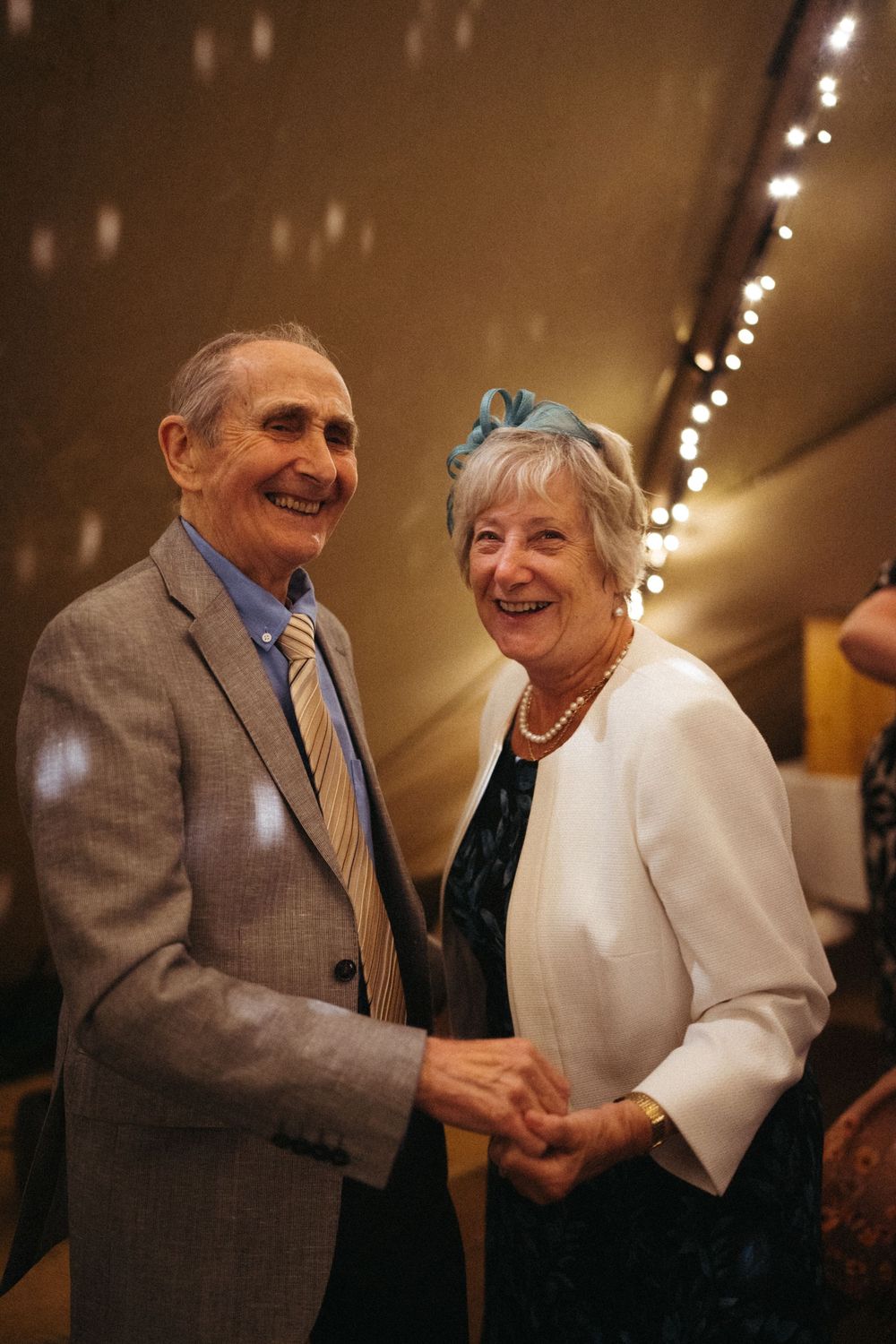 An elderly couple share a joyful moment together at an indoor celebration with twinkling lights in the background.