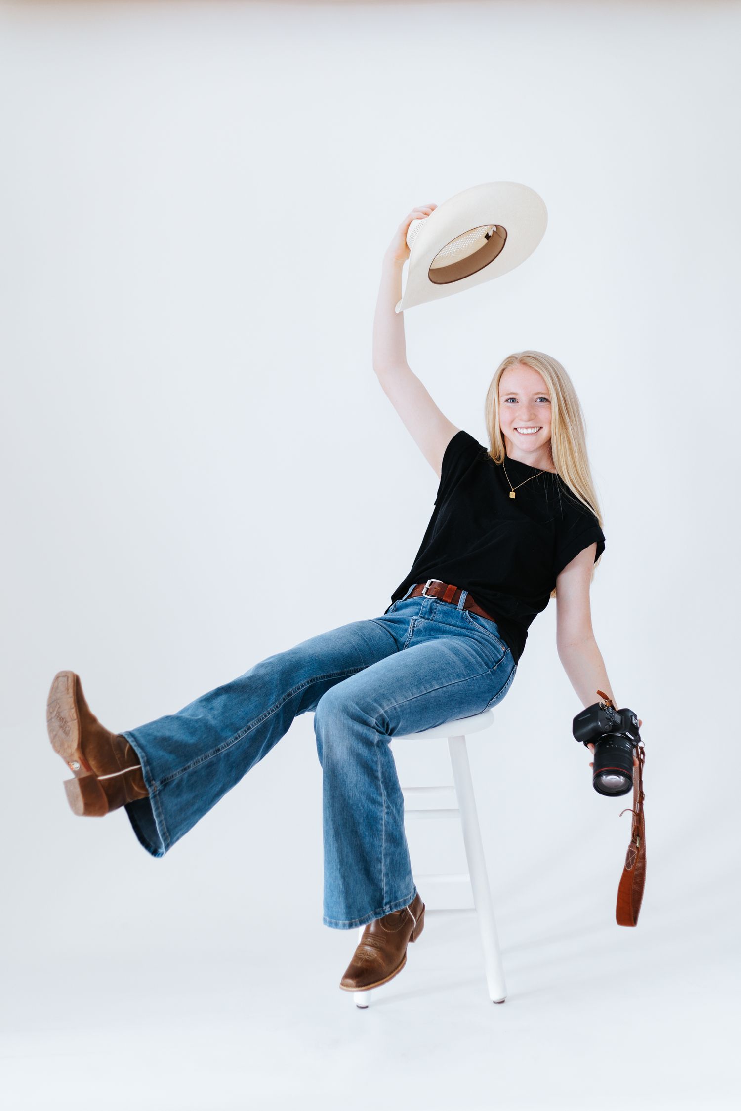 A photographer in western wear poses playfully on a white stool with a camera.