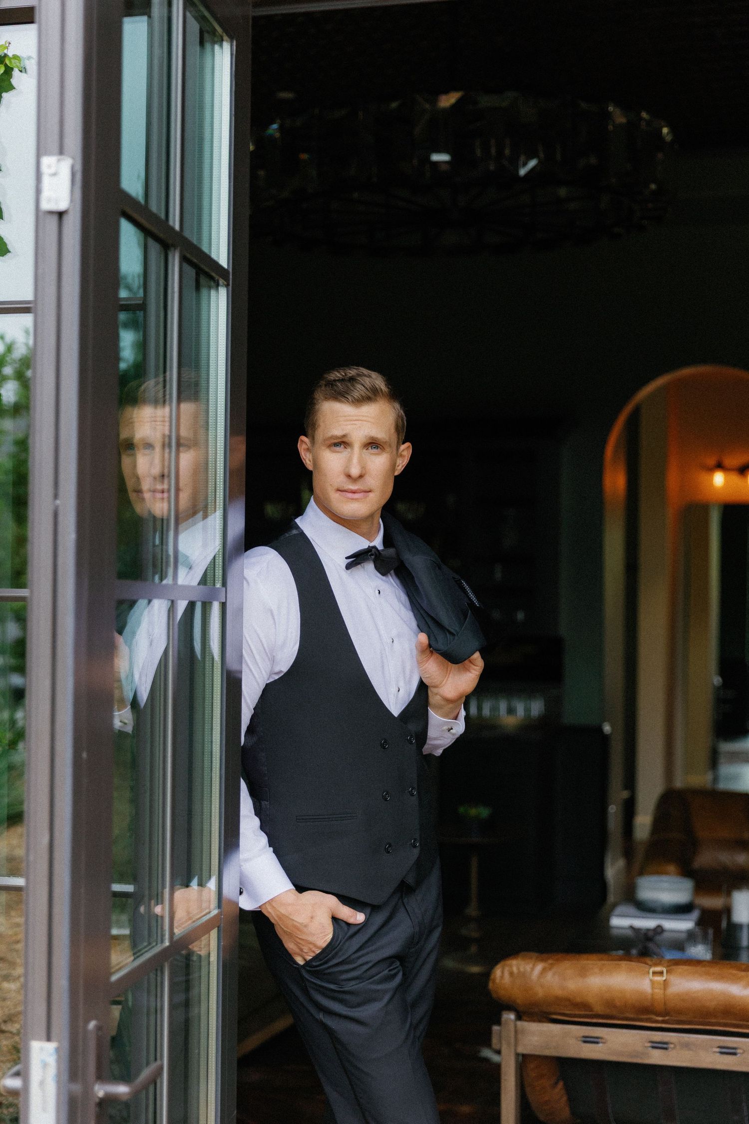 Well-dressed server in formal vest and bowtie stands confidently by glass doorway of upscale restaurant.