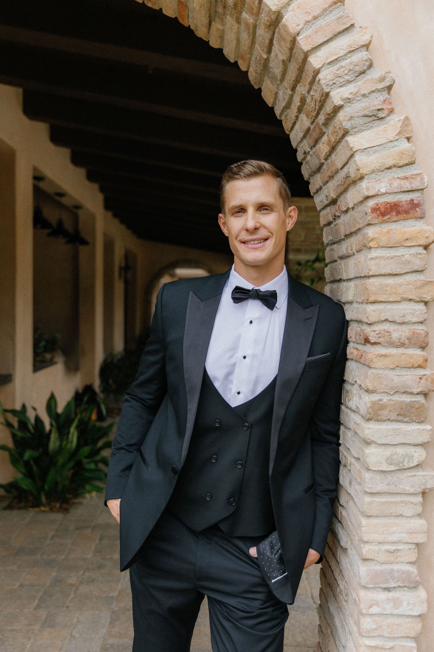 A person in a classic black tuxedo with bow tie poses in an elegant archway with exposed brick details.