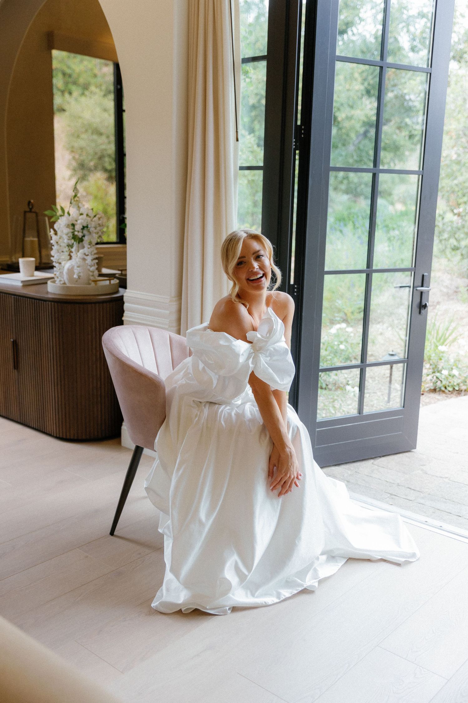 A person in an elegant white wedding gown sits gracefully in a pink chair near French doors in a sunlit room.