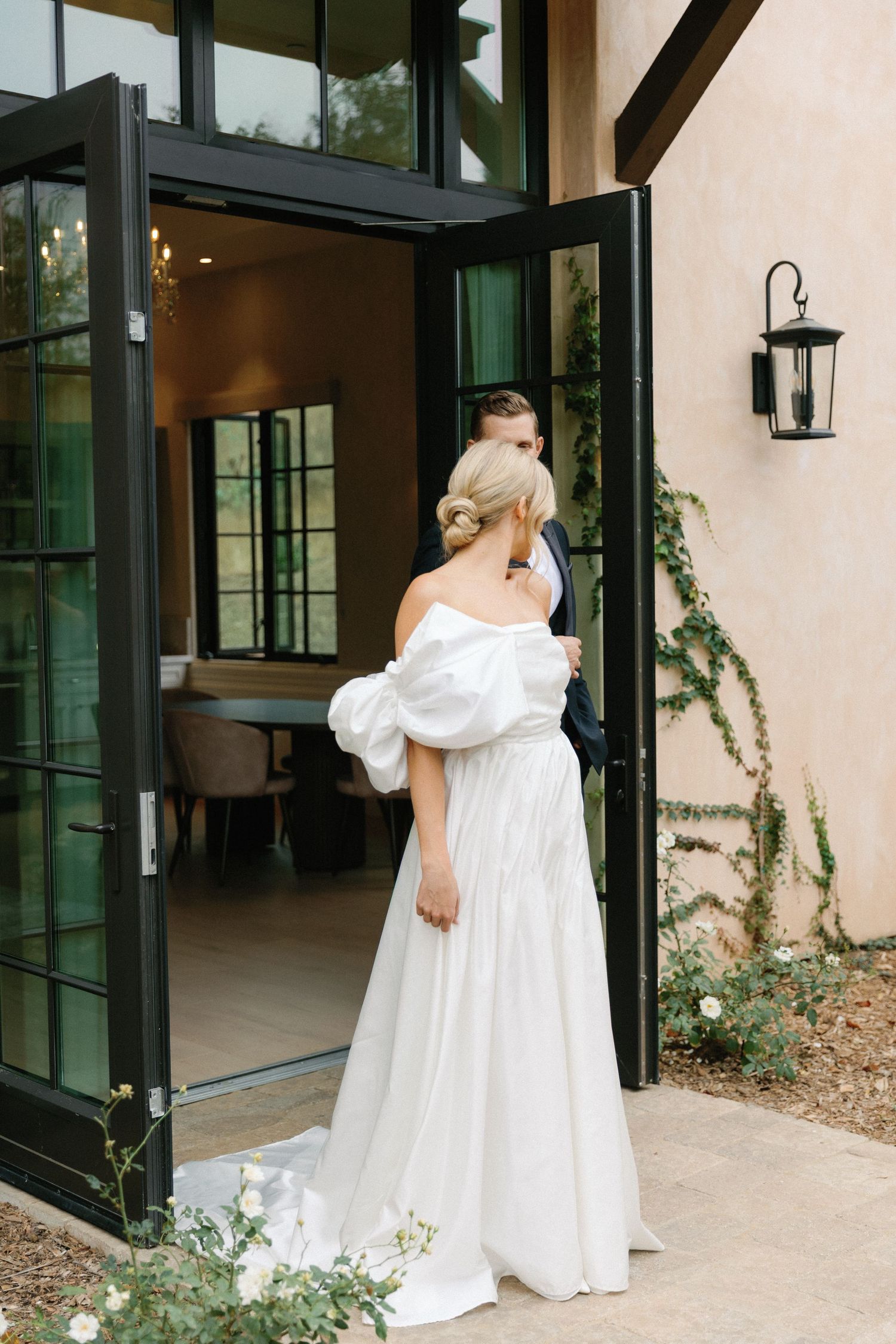 A bride in an elegant off-shoulder white gown stands by glass doors of a venue with exterior wall lantern.