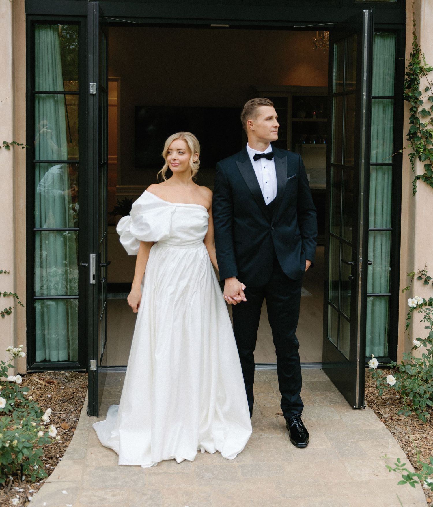 A bride in an off-shoulder white gown stands with a groom in black tuxedo outside elegant French doors.
