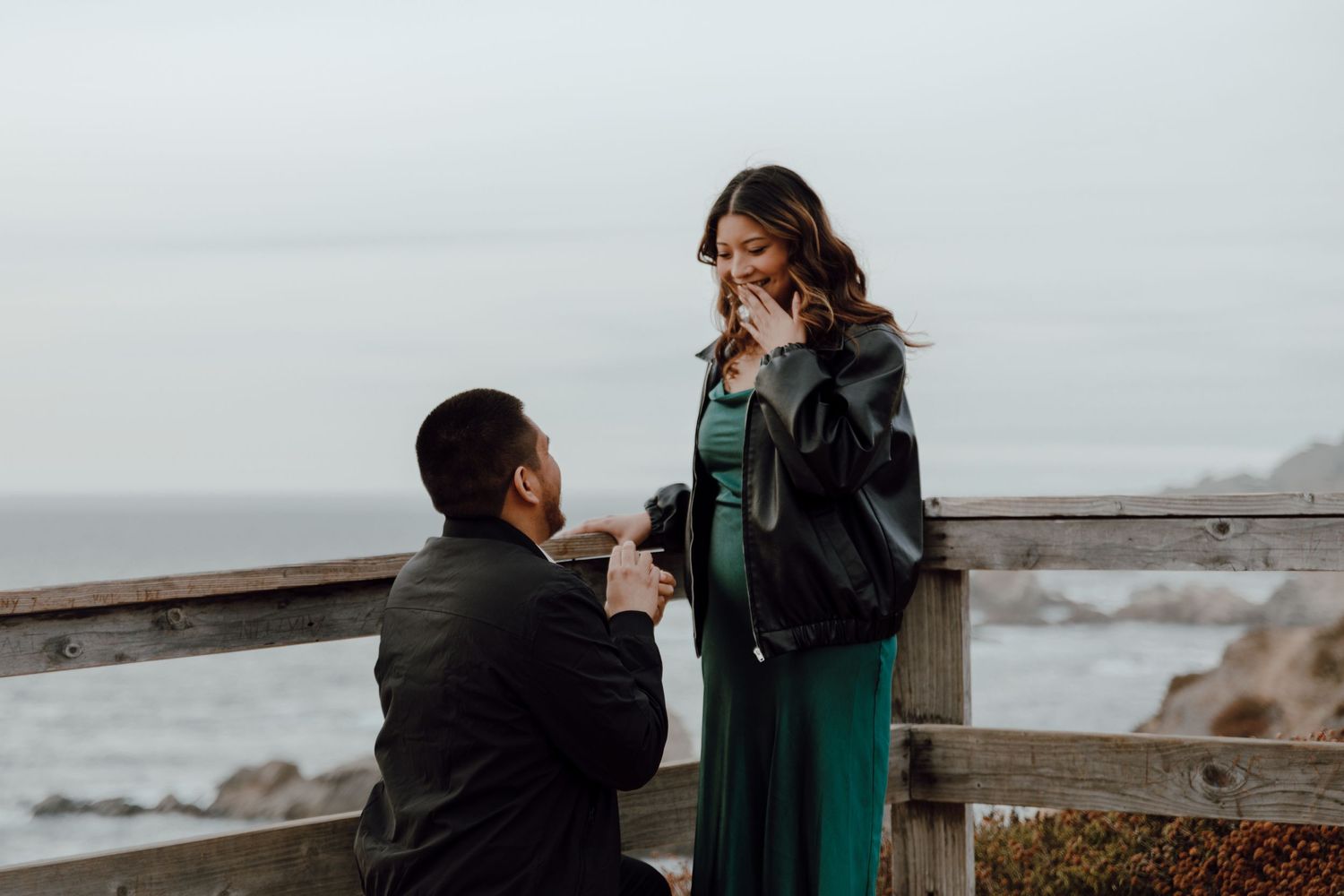 A romantic proposal moment takes place on a wooden deck overlooking a misty coastal landscape on a cloudy day.