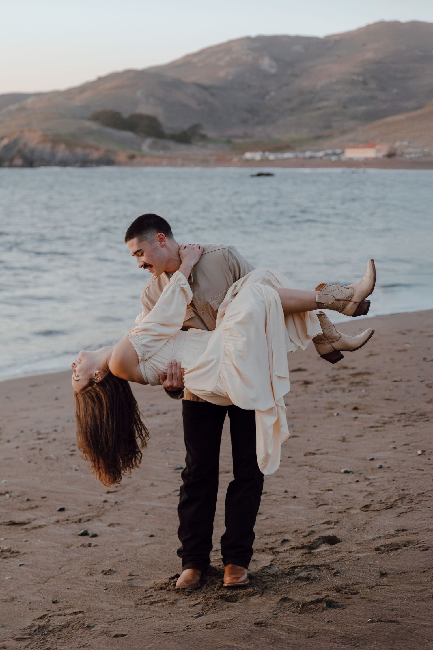Romantic couple embraces in a dramatic dip pose on a sandy beach at sunset with mountains in the background.