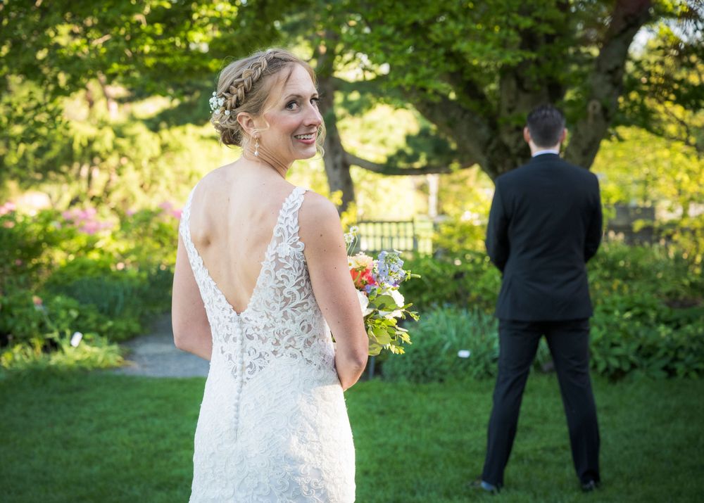 Back view of bride in lace dress with bouquet as groom stands in background among garden greenery.