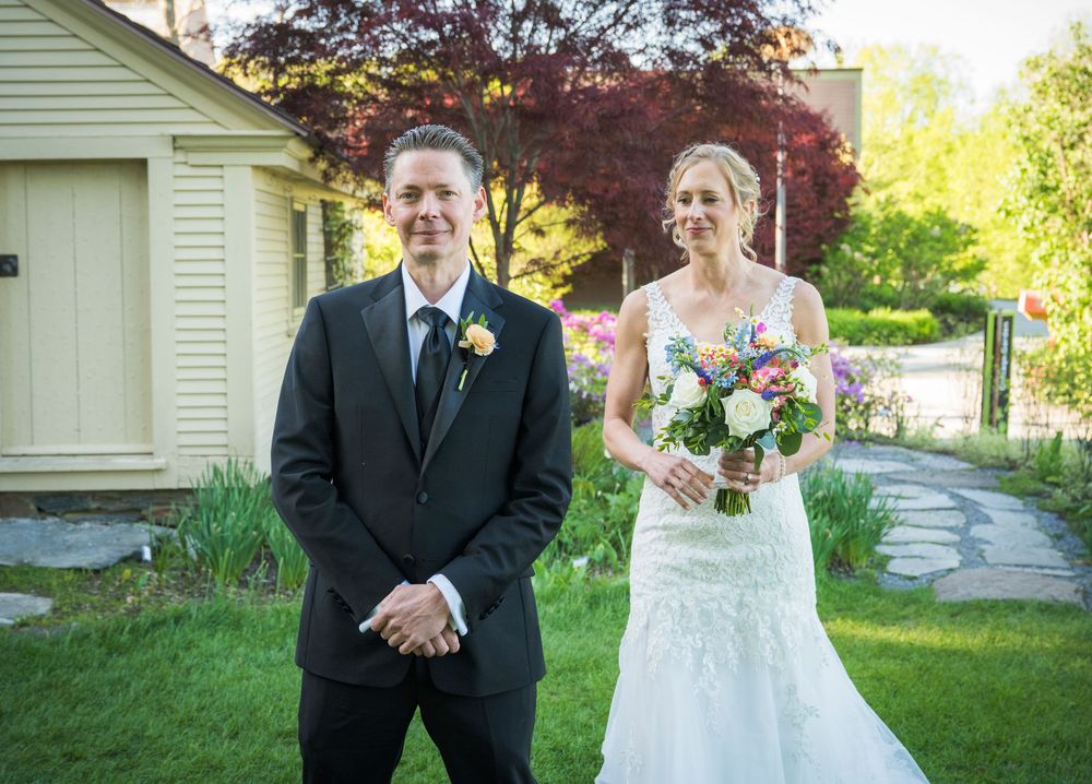 A couple in wedding attire stands outside a white house with spring gardens and red maple trees.