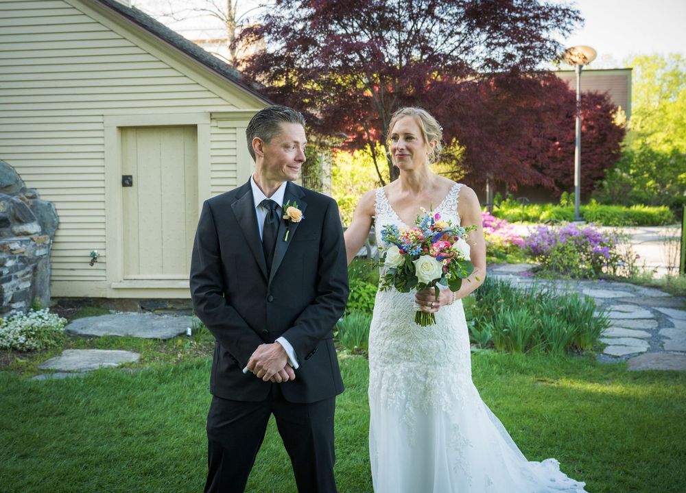A wedding couple poses together on a lawn in front of a white building with spring foliage.