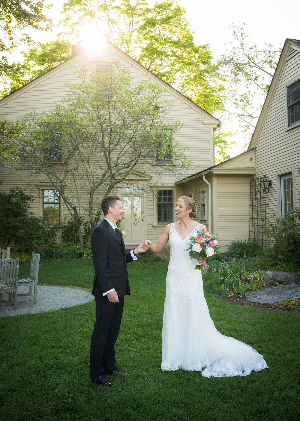 A wedding couple shares a moment on a lawn with white colonial houses and sunlit trees in the background.