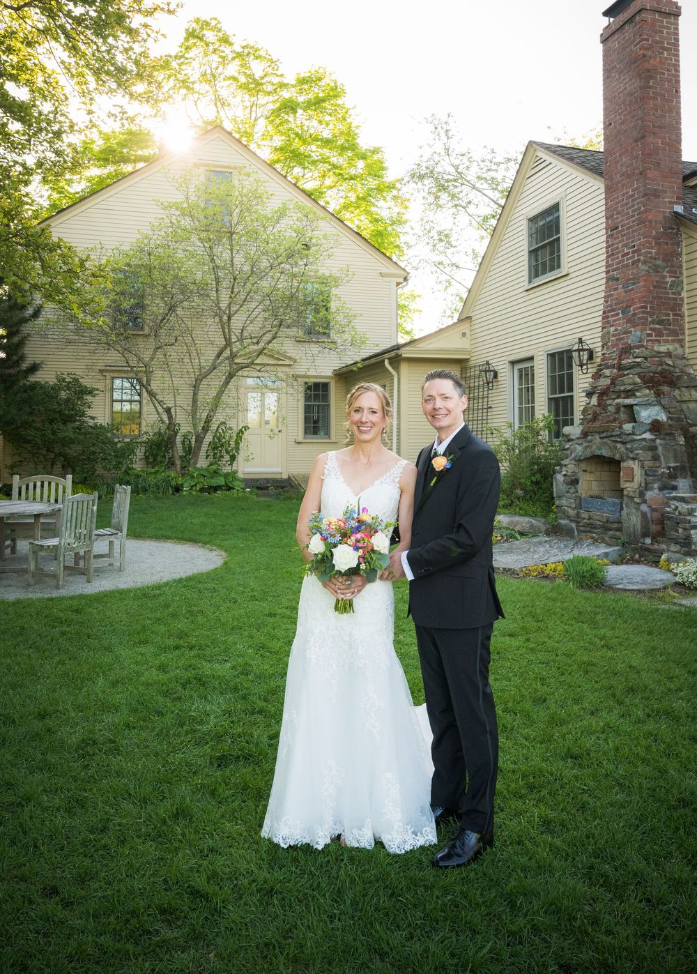 A newlywed couple poses for a portrait in front of a traditional house with a brick chimney.