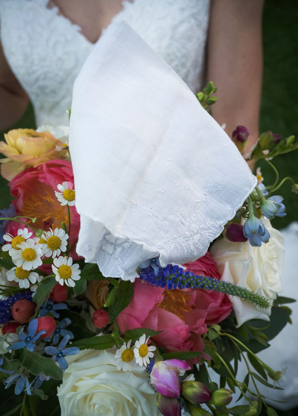 A colorful bridal bouquet with roses, daisies and blue flowers next to a white lace handkerchief.
