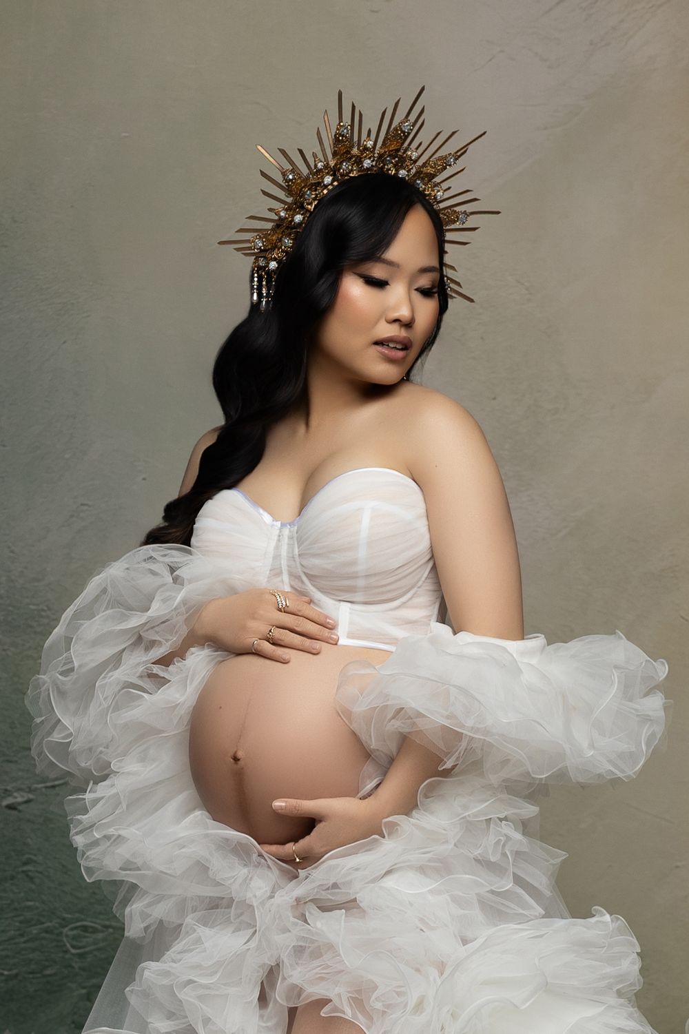 A maternity photoshoot featuring a delicate white tulle dress and golden spiked crown headpiece against a textured backdrop.