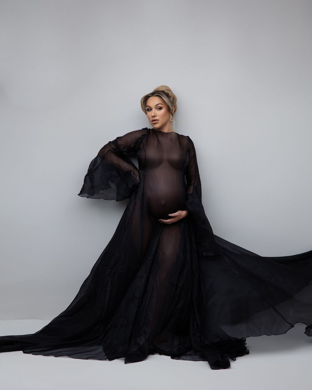 An elegant maternity portrait with a flowing black sheer gown and cape against a light gray studio backdrop.
