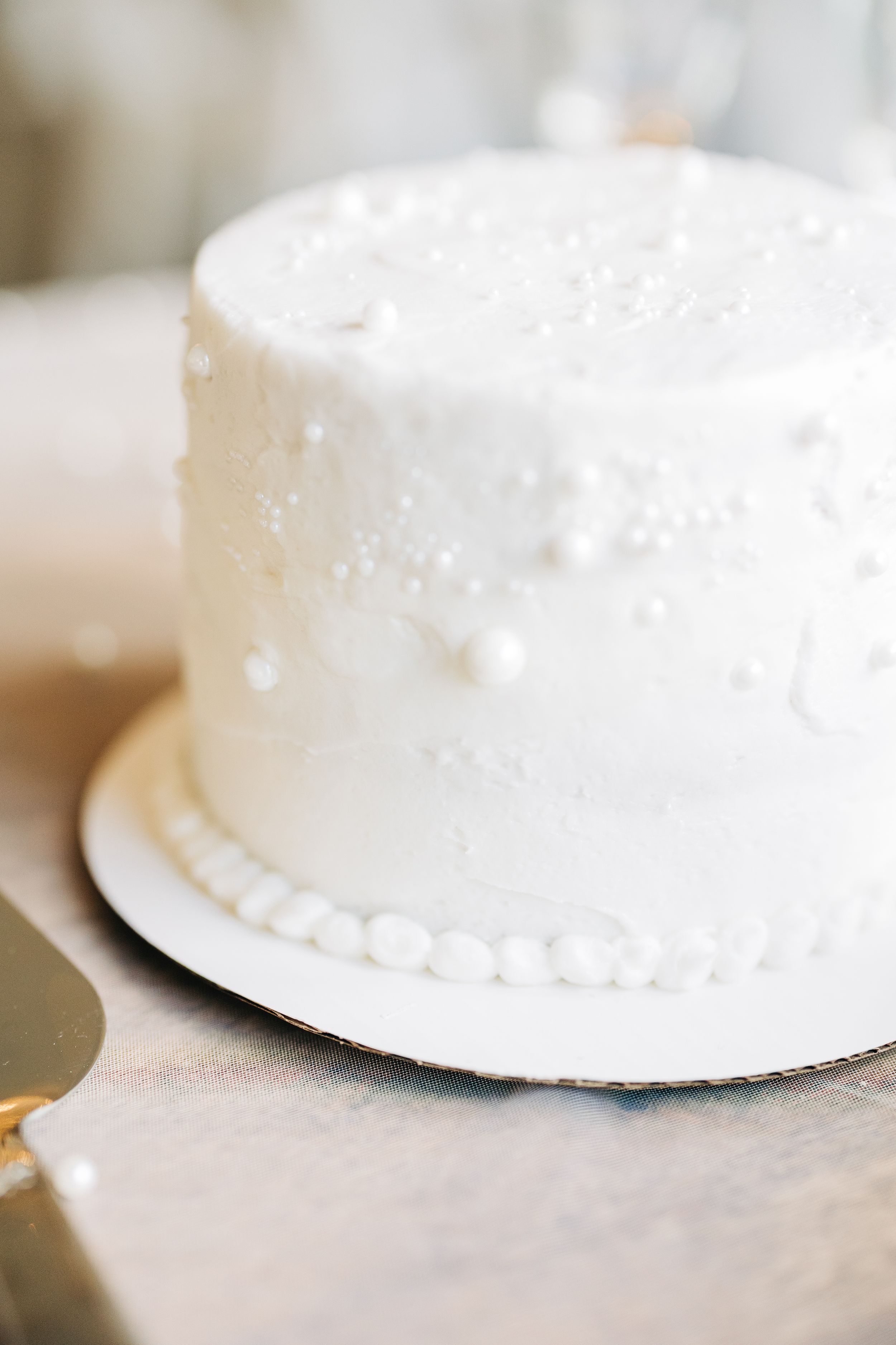 A simple white frosted cake with delicate pearl details along the base displayed on a metallic surface