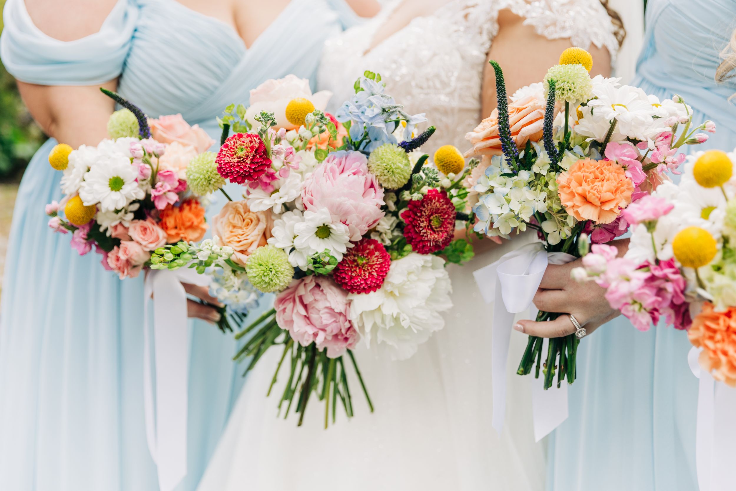 Bridesmaids in light blue dresses hold vibrant bouquets of pink, yellow and white flowers at a sunny outdoor wedding.