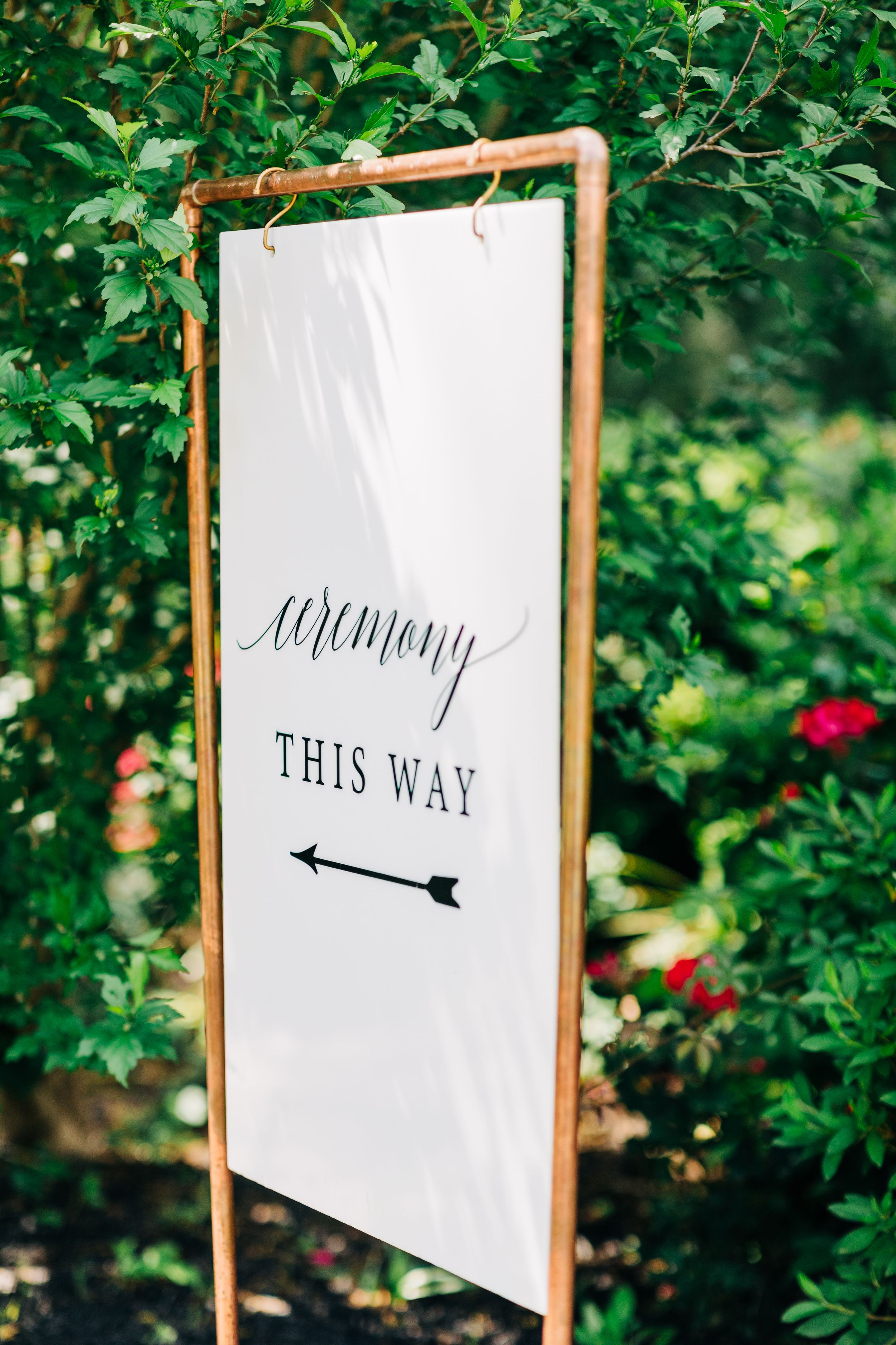 A wedding sign with "ceremony this way" text and arrow mounted on copper pipes among green foliage in a garden setting at the Wildflower 301