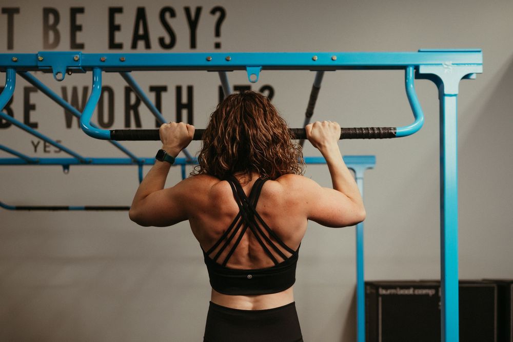 Athlete in black workout attire performs pull-ups on blue gym equipment against grey wall background.