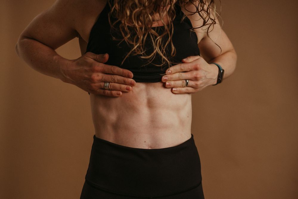 Person wearing black athletic wear shows defined abdominal muscles against brown background in fitness portrait.