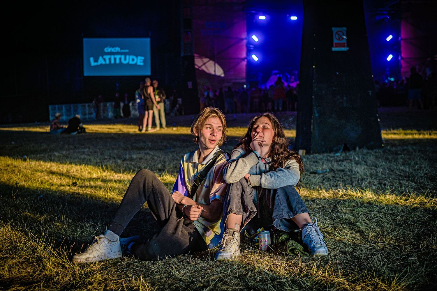 Two festival-goers sitting on grass at night with blue stage lights illuminating the background.