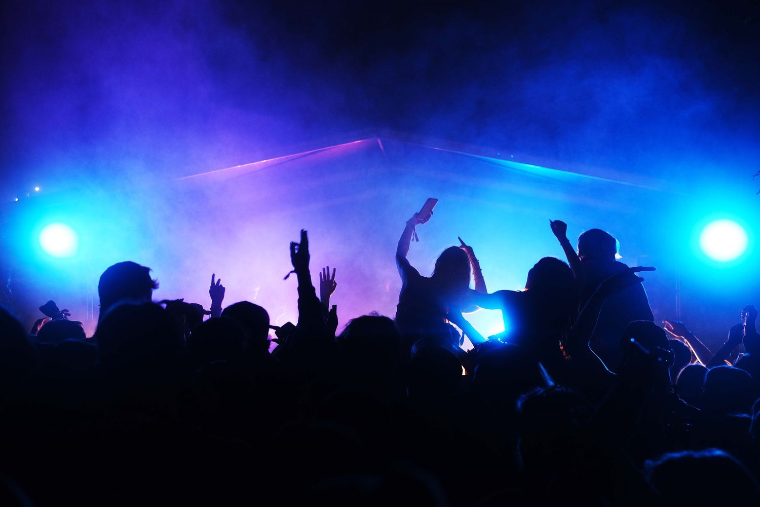 Silhouettes of concert crowd with raised hands against vibrant blue stage lighting and laser beams.