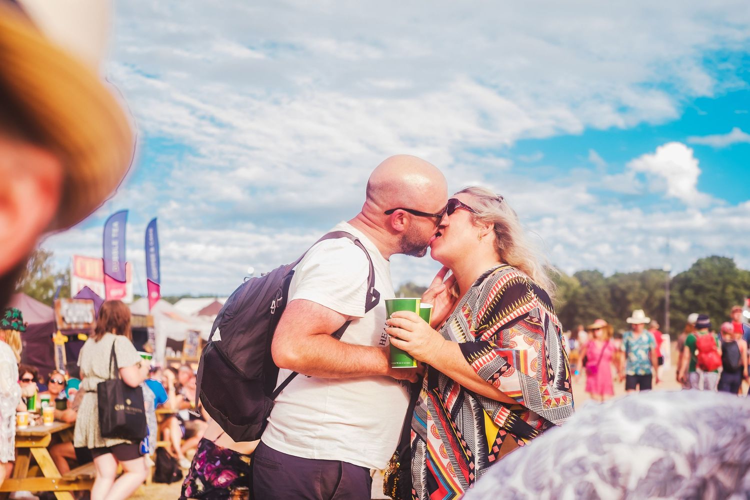 A couple shares a kiss at an outdoor summer festival with blue sky and colorful flags visible in the background.