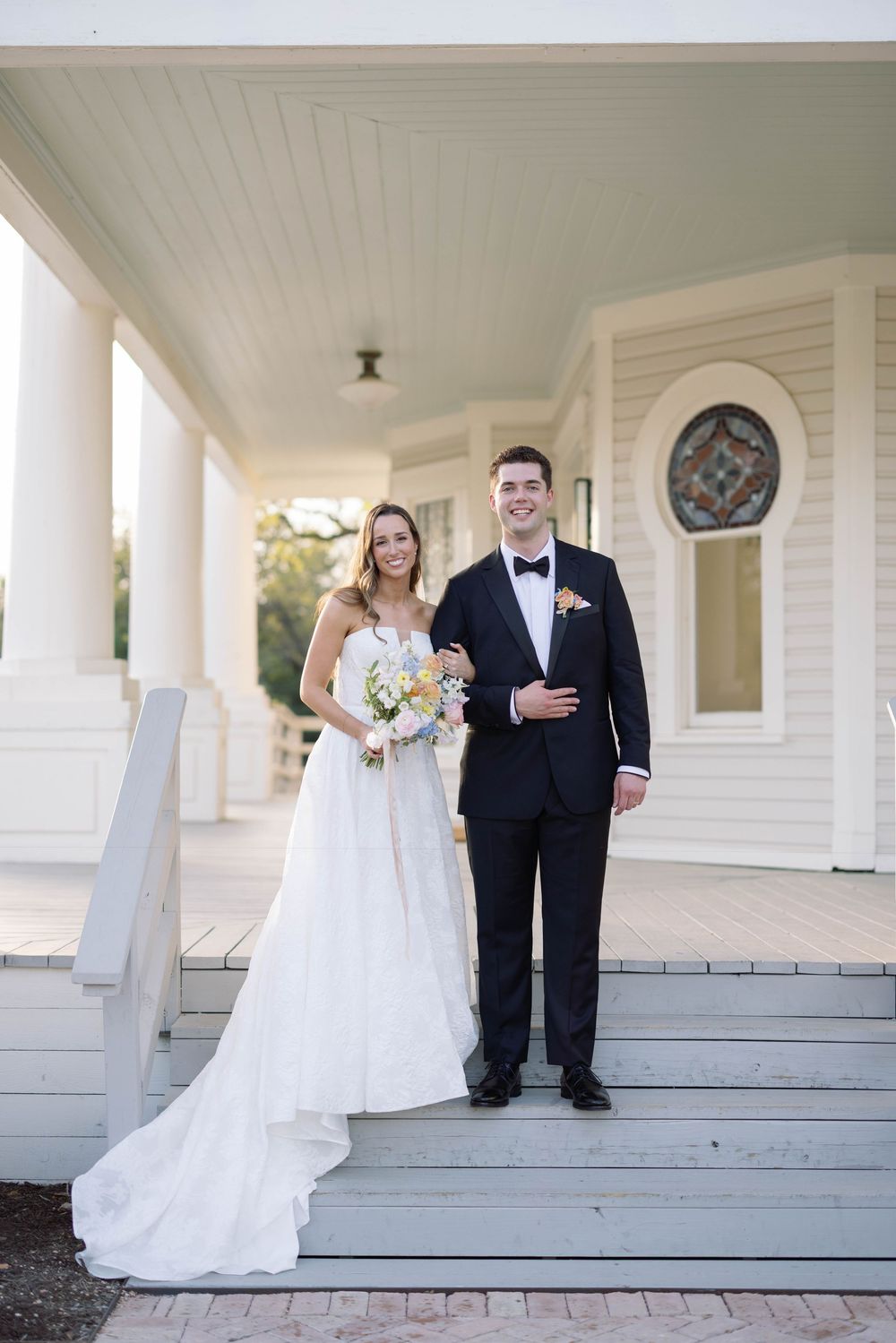 Wedding couple stands on a classic white porch with pillars in romantic photo series.