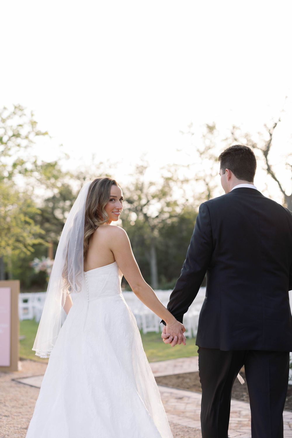 Bride and groom walking hand in hand along outdoor path with flowing white dress and veil.