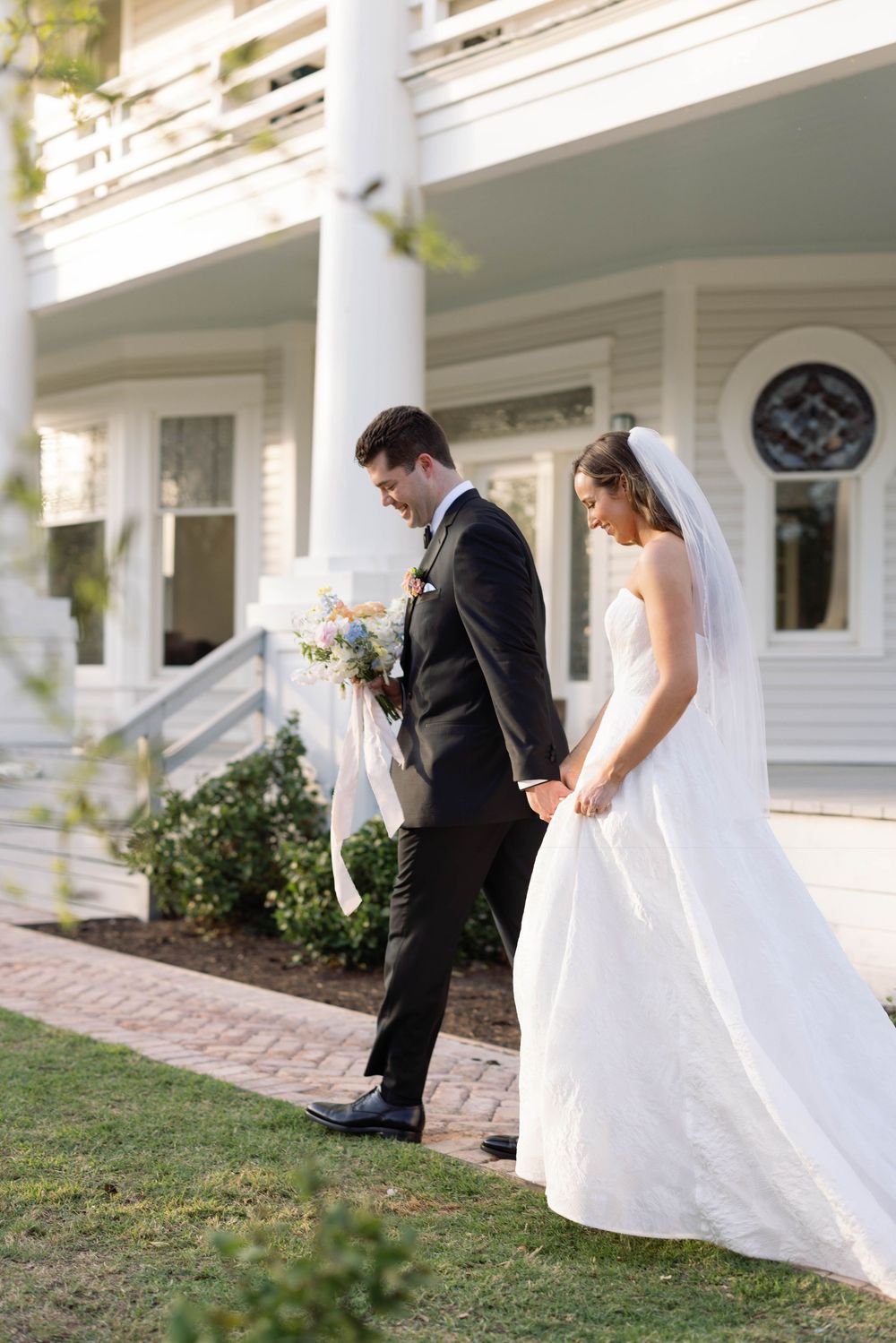 A couple in wedding attire walks along a brick path beside a white pillared house surrounded by gardens.
