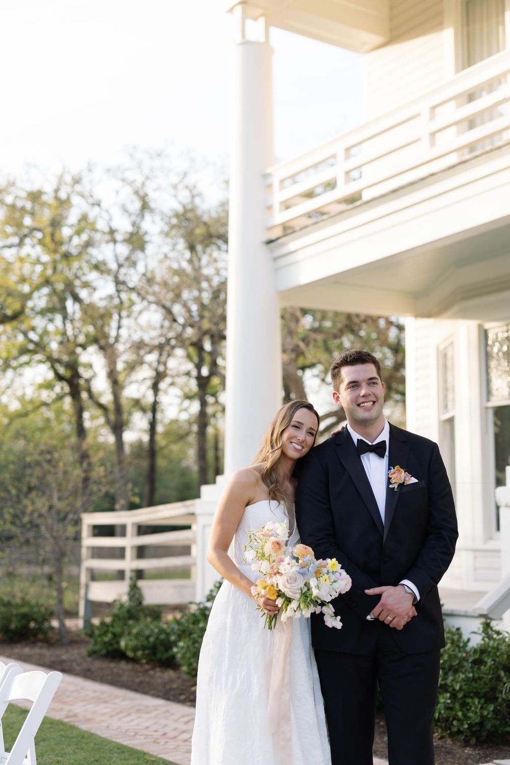 Newlyweds share a joyful moment on a white columned porch with the bride holding a pastel flower bouquet.