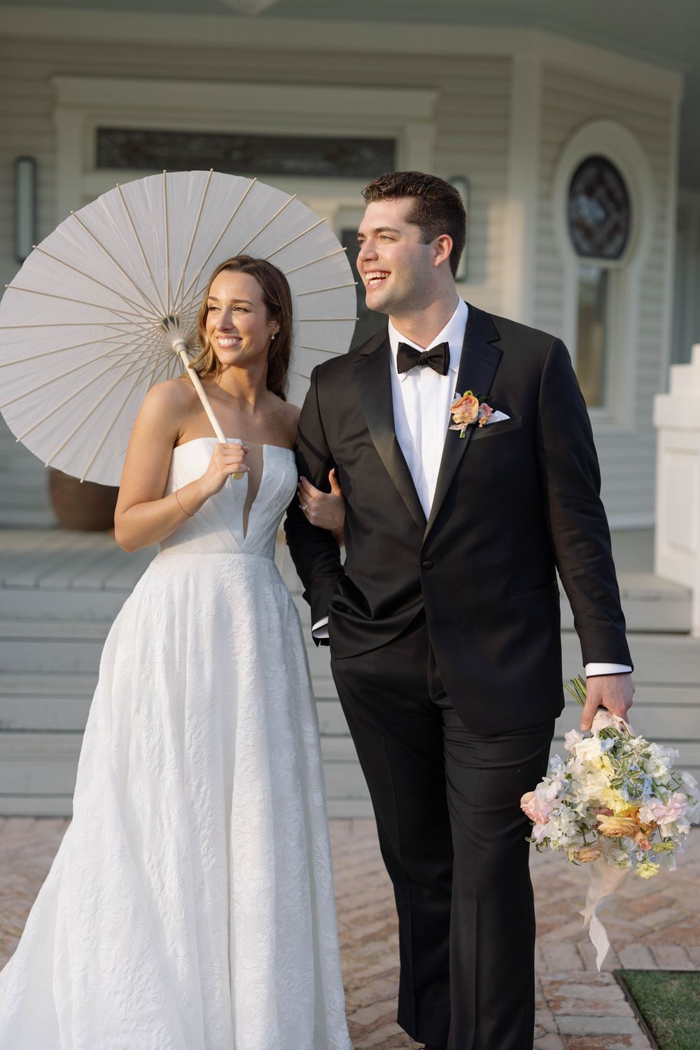 A bride holds a white parasol and laughs joyfully in a white gown next to a groom in a classic black tuxedo with boutonniere.