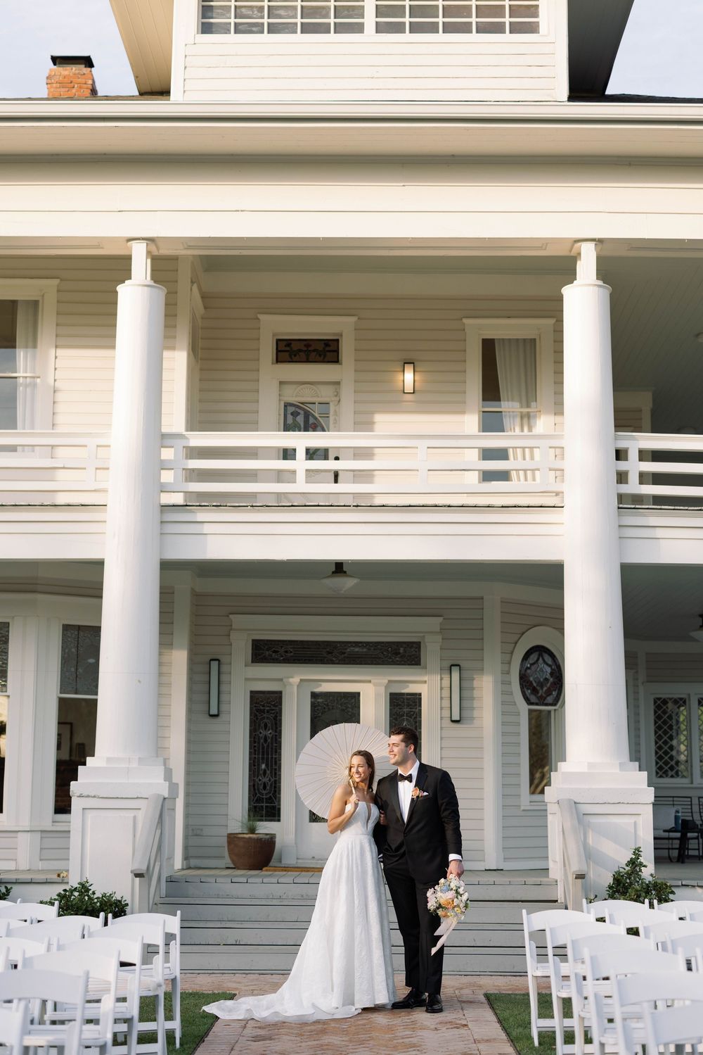 A wedding couple stands on a grand white porch with columns, with rows of white ceremony chairs set up on the lawn.