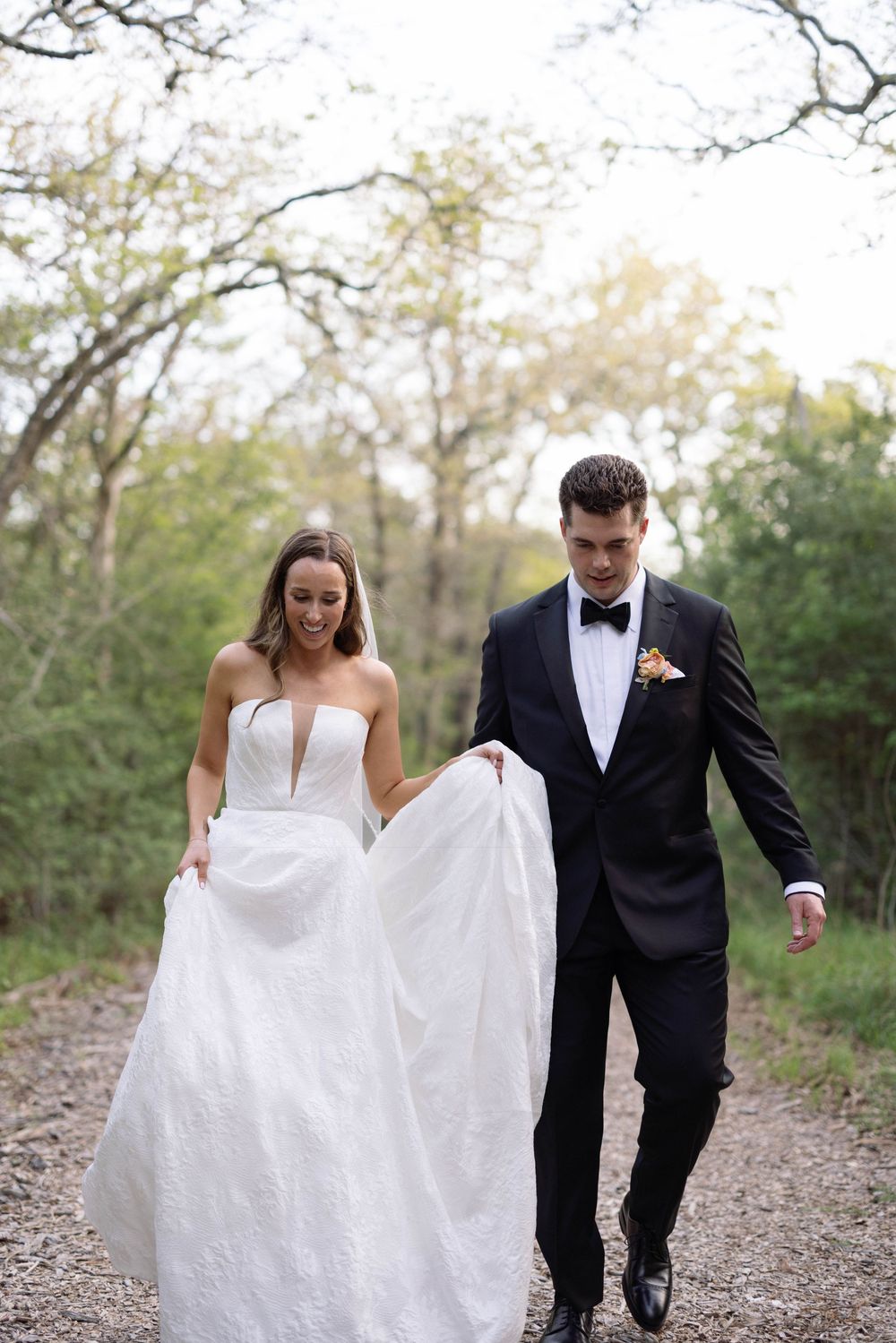 A couple walks together on a gravel path with the bride holding her wedding dress skirt.