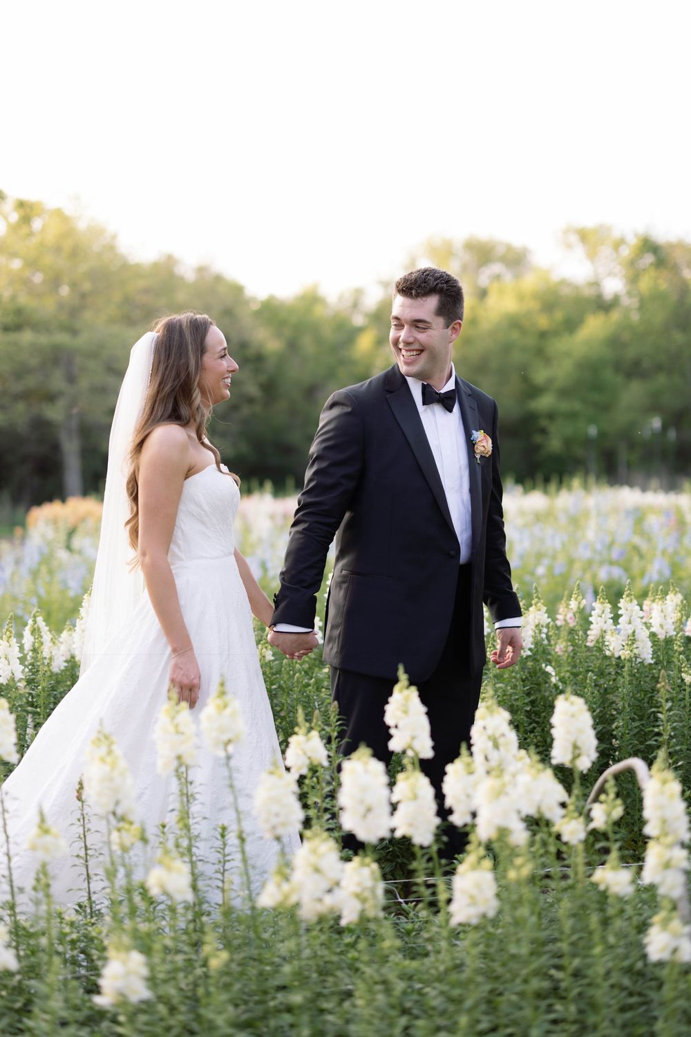 A bride and groom share a candid moment while walking through a garden filled with white blooming flowers.