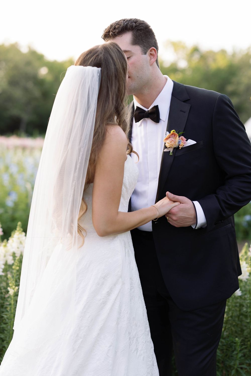 A wedding couple shares an intimate moment in a flower-filled garden, with the bride in white and groom in black tuxedo.