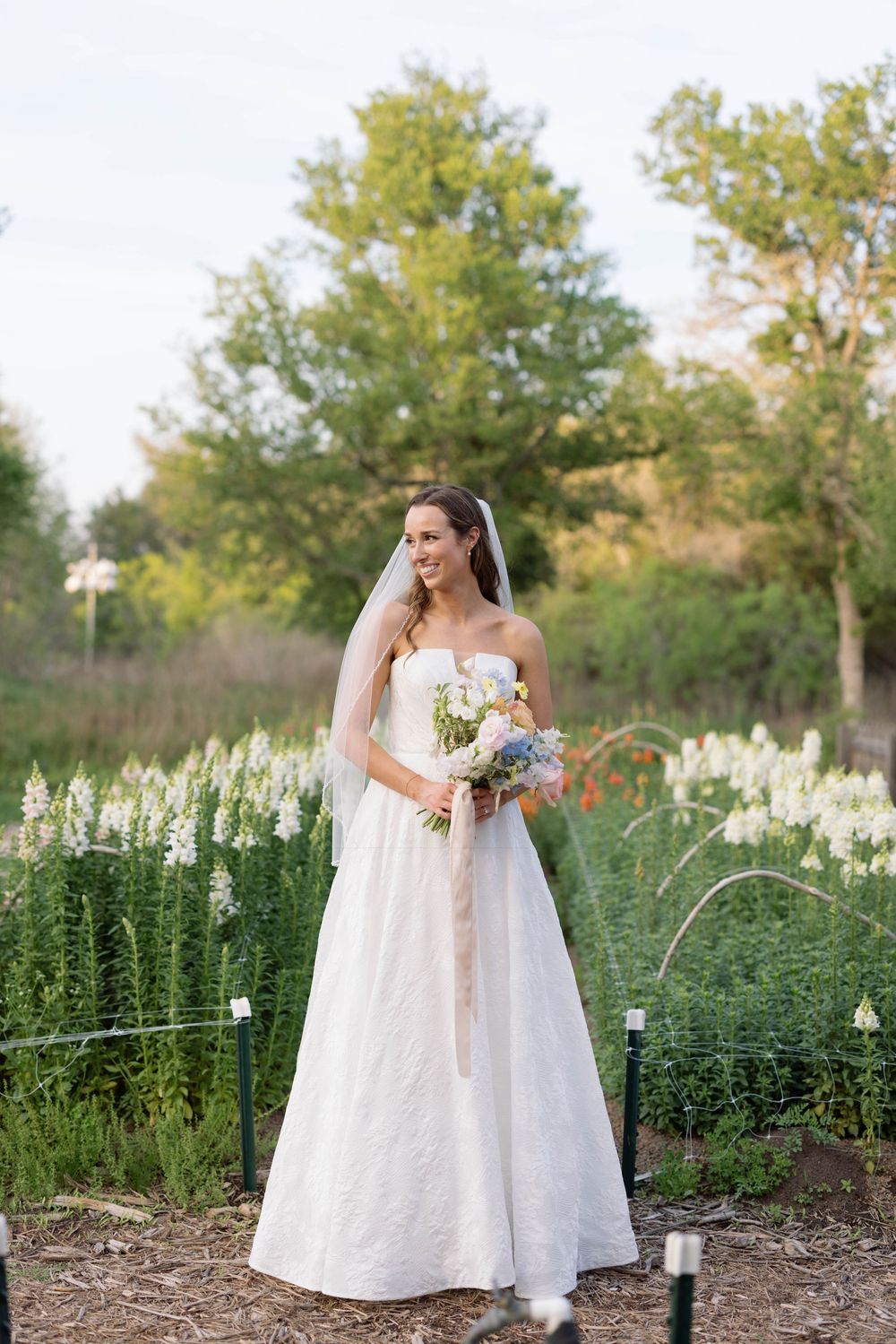 Classic white wedding gown with long veil framed by garden vegetation and pathway lights at sunset in natural setting.
