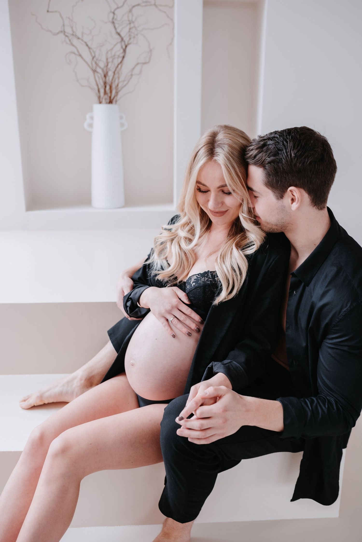 A romantic maternity photoshoot shows an intimate moment in a minimalist white room with a decorative vase.