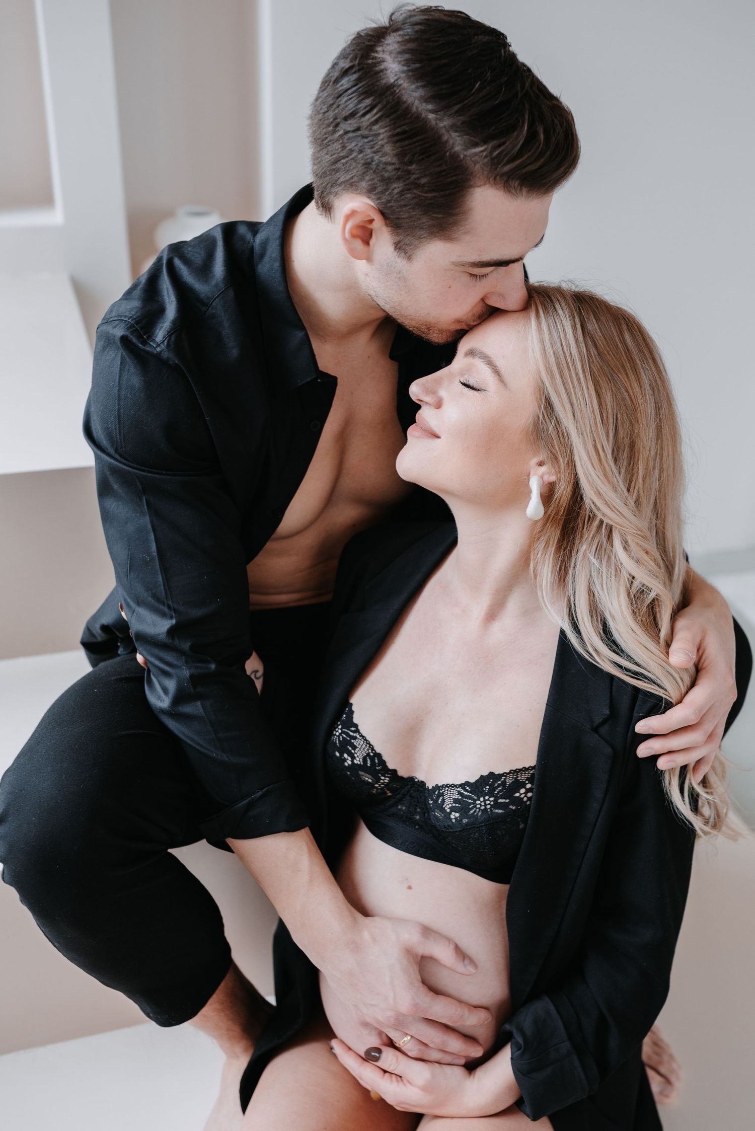 A romantic couple in black formal wear embrace intimately in a photo series with soft lighting and neutral background.