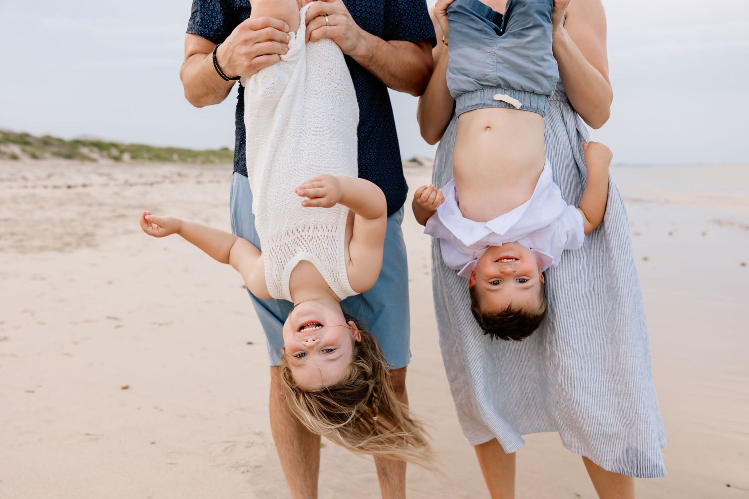 Two children held upside down by adults while playing and laughing at the beach on a sunny day.