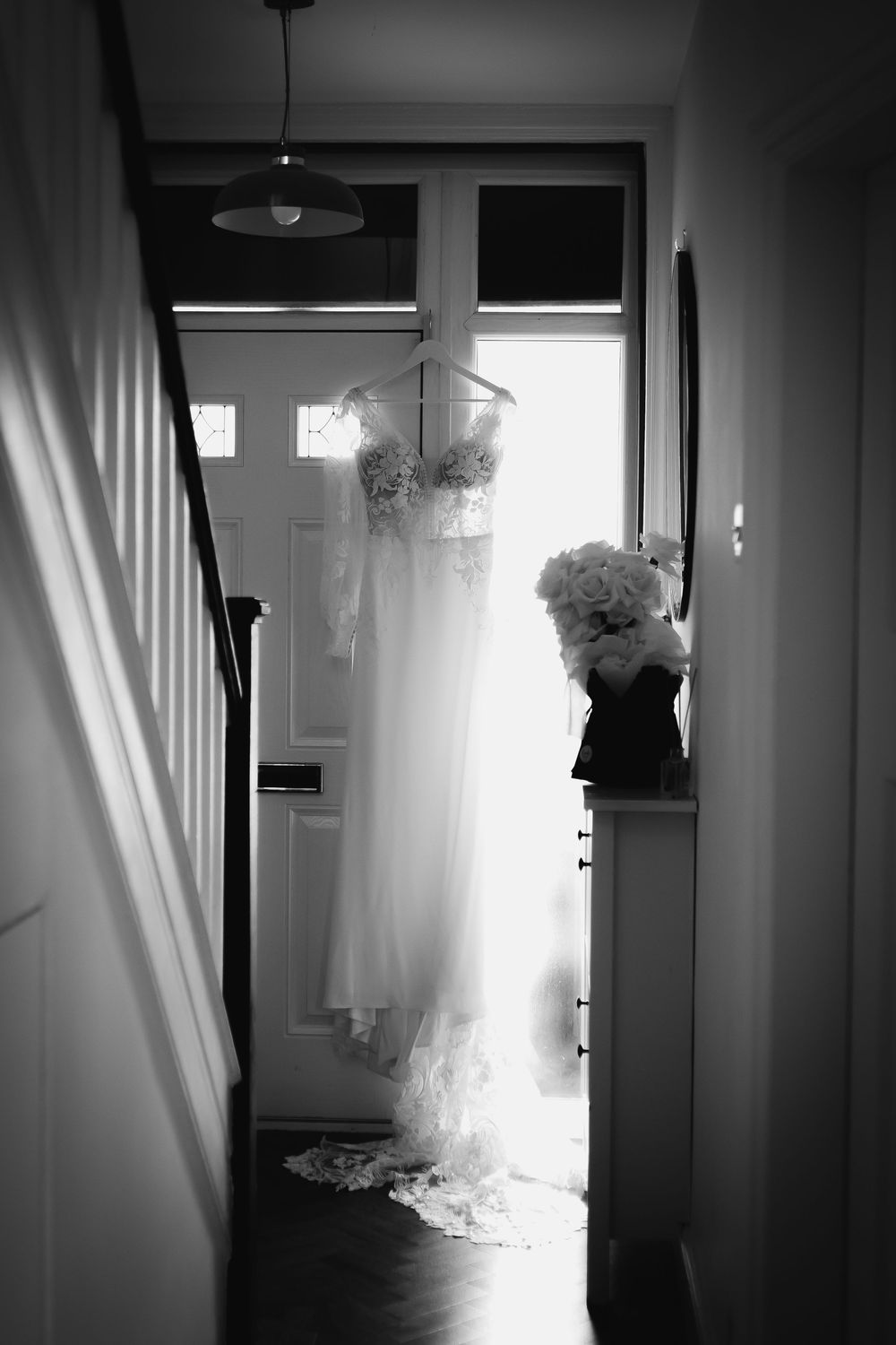 Wedding dress hanging in doorway with dramatic natural backlighting creating ethereal silhouette.