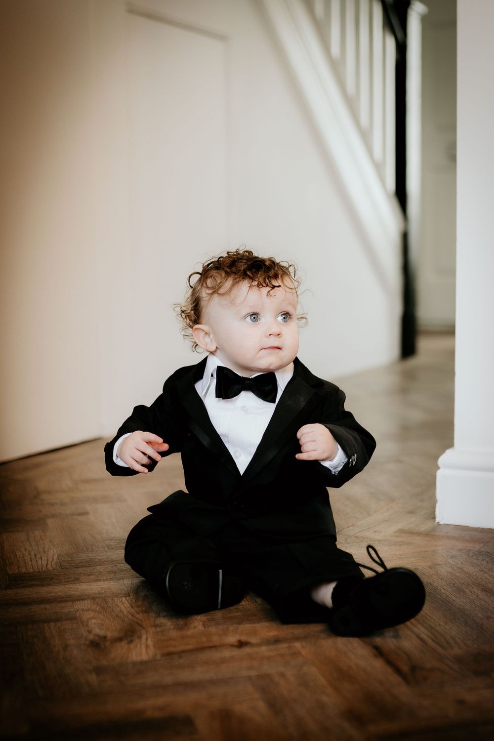 A toddler in a formal black tuxedo and bowtie sits on hardwood flooring near white stairs.
