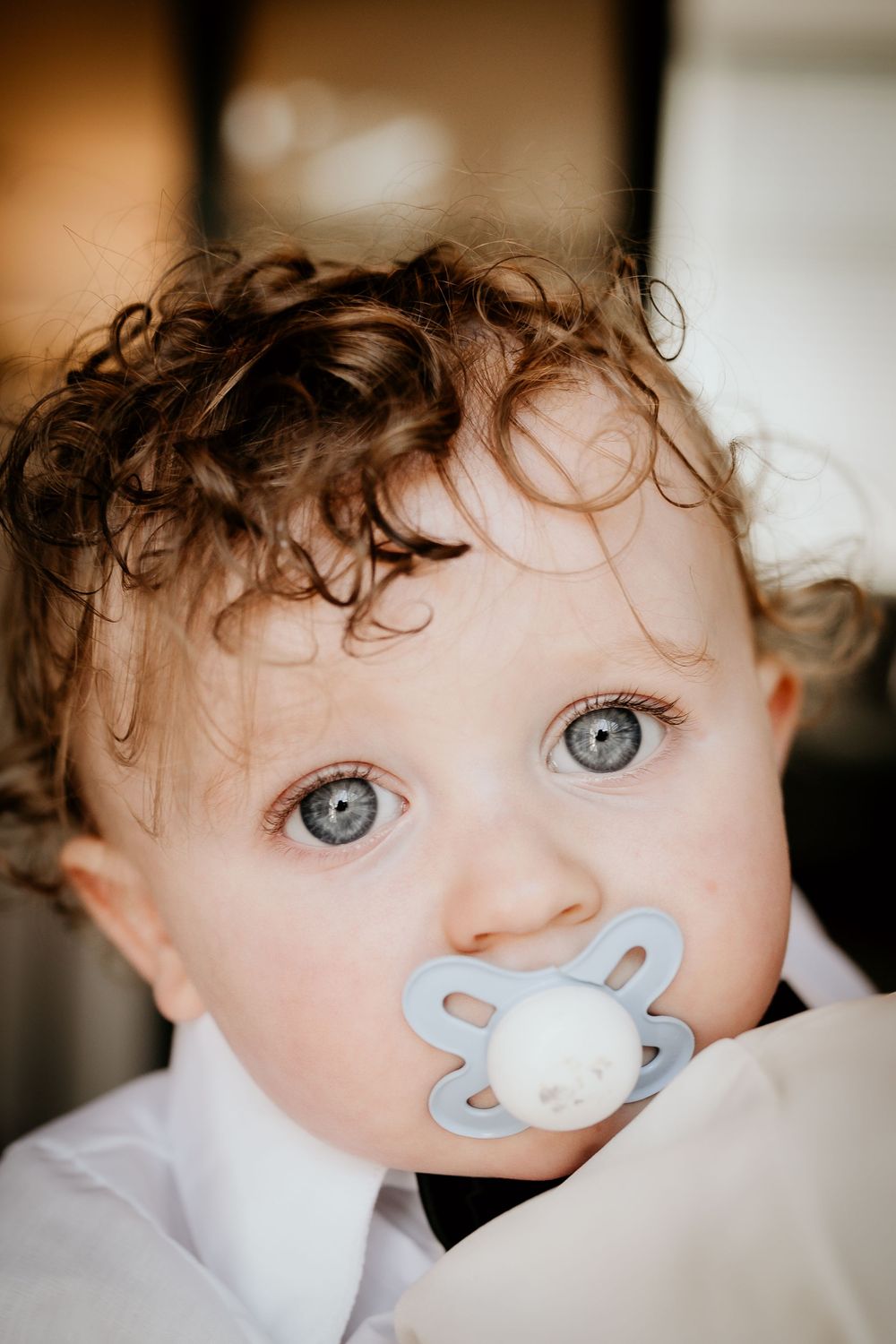 Close-up view of a baby with bright blue eyes and curly hair wearing a light-colored pacifier.
