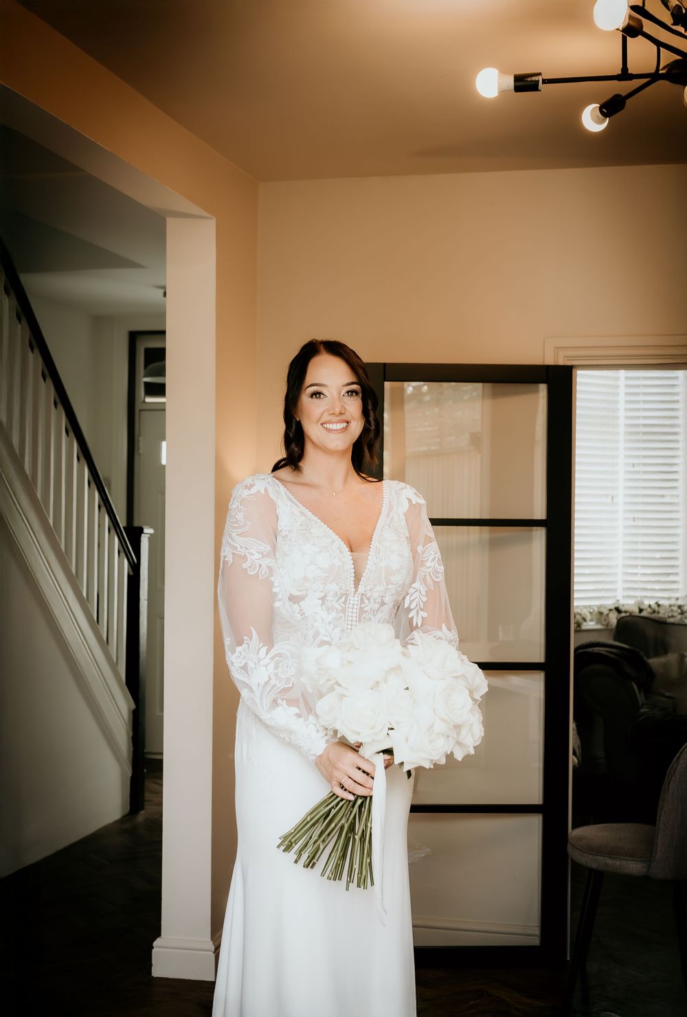 A bride in an elegant white lace dress holds a bouquet while standing in a home interior.