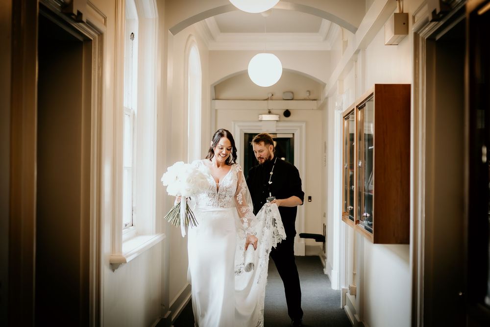 A wedding couple walks down a sun-lit hallway with white walls and glowing globe pendant lights creating a romantic ambiance.