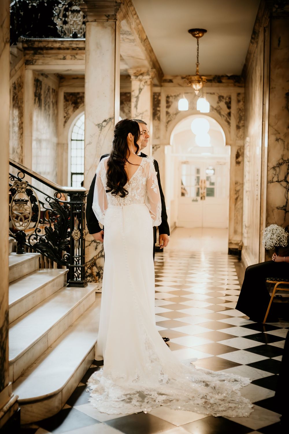 Elegant white lace wedding gown photographed in an ornate hallway with checkered floor and arched windows.