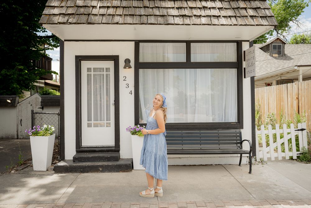 A quaint cottage-style house with cedar shingles, black trim, and white picket fence featuring someone in a blue dress out front.