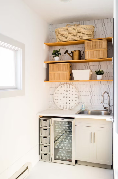 Modern white laundry room with wooden shelving, storage baskets, and white subway tile backsplash against bright windows.