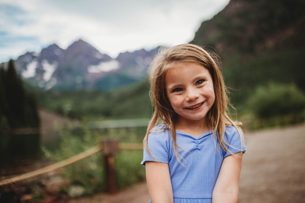 Young girl in blue shirt smiles while sitting outdoors with mountain landscape in background.