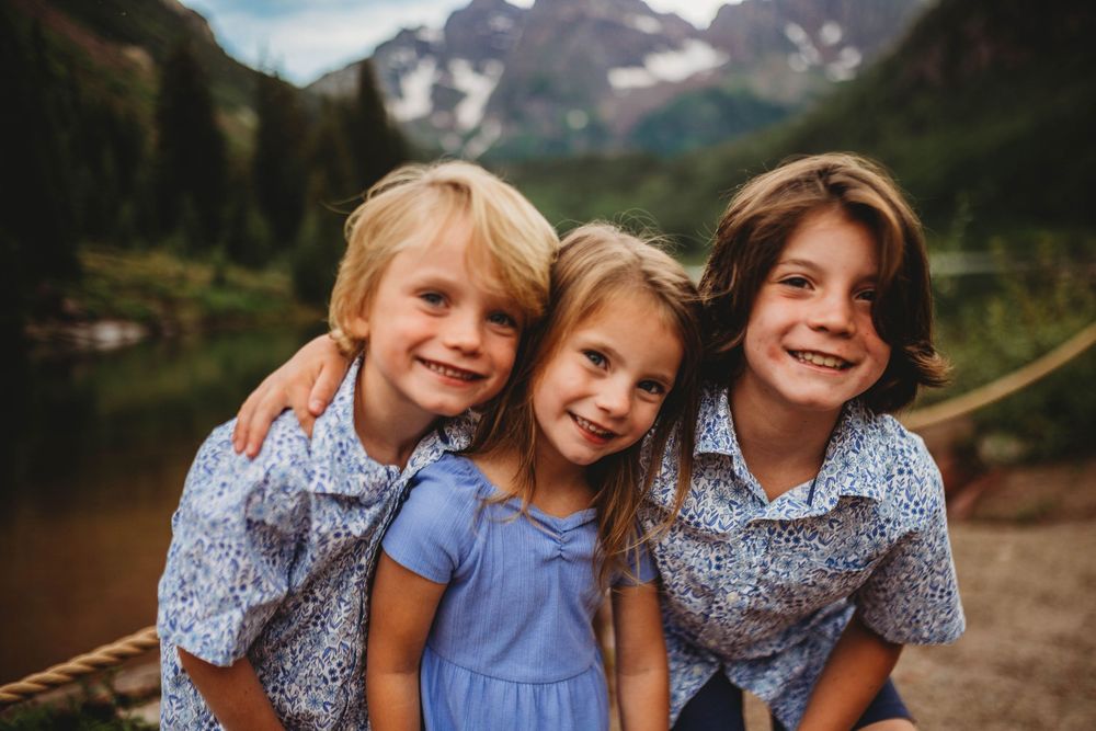 Three children smile together against a scenic mountain lake backdrop with evergreen forest and snowy peaks.